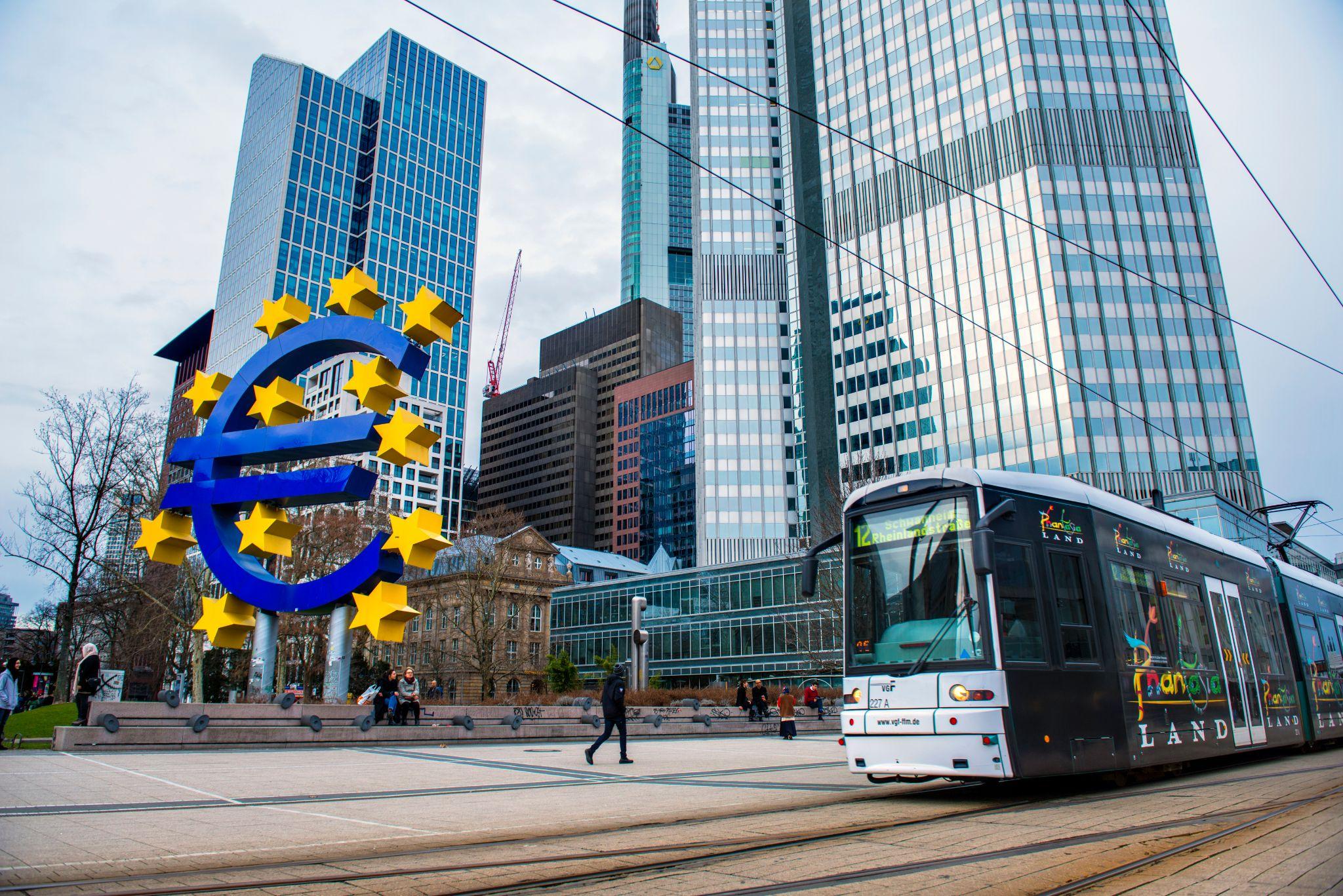 A modern tram passes by a large Euro symbol sculpture in a financial district with high-rise buildings in the background.