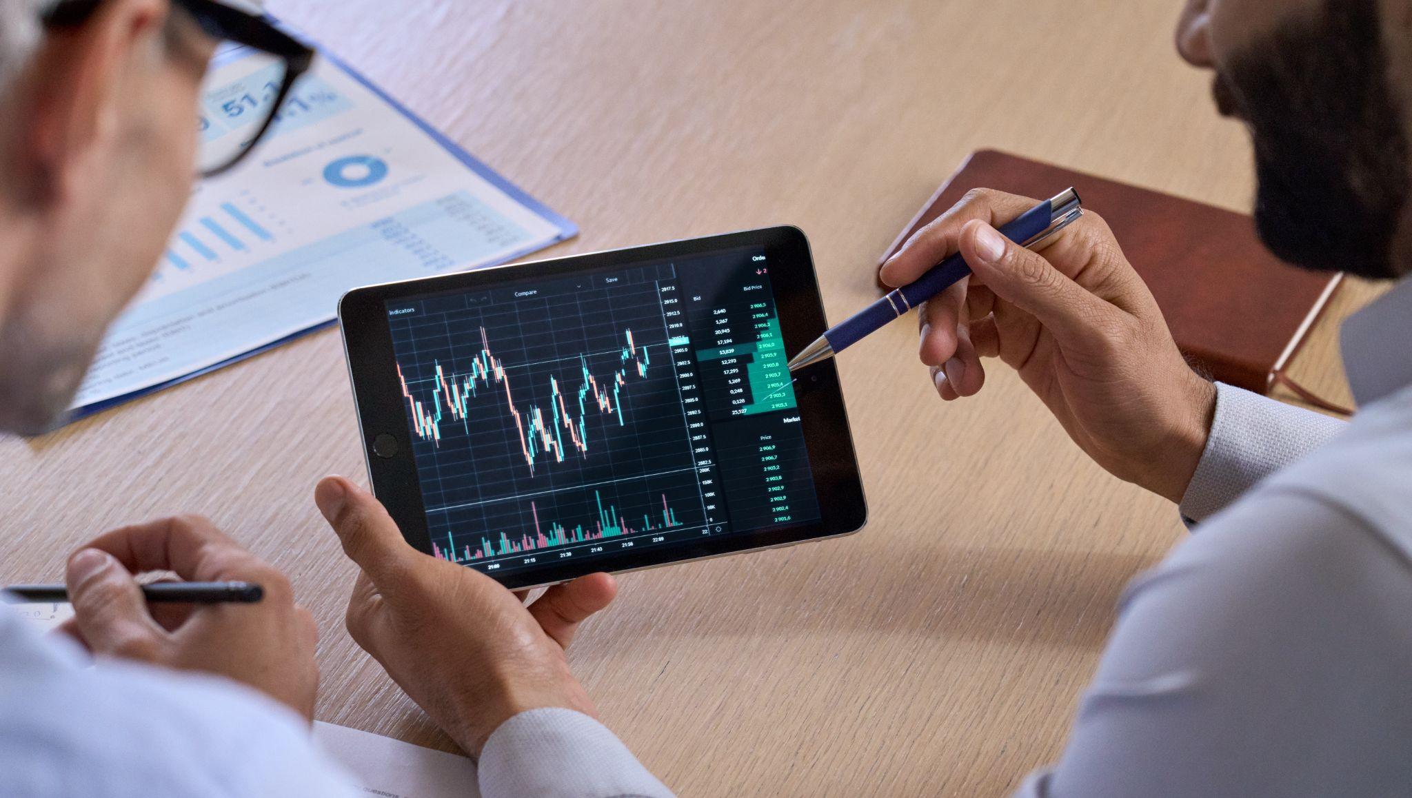 Trader showing crypto chart to investor on tablet, analyzing market, discussing risks and profit. Over-shoulder view.