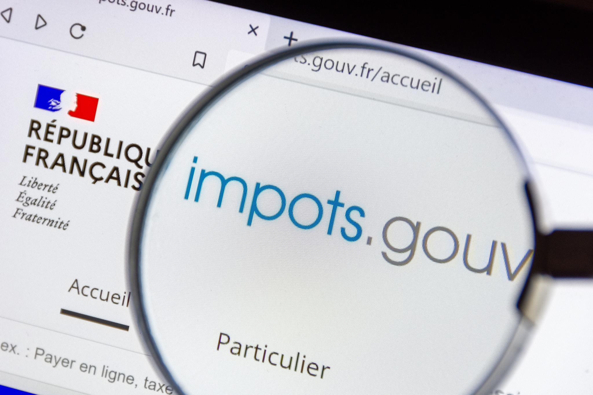 Magnifying glass highlighting the 'impots.gouv' website on a computer screen with the French Republic logo visible.