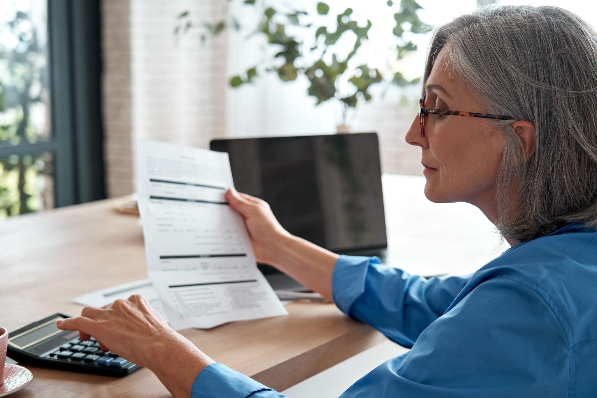 Senior woman managing finances: holding bill, using calculator, budgeting, planning loan debt and pension at home office.