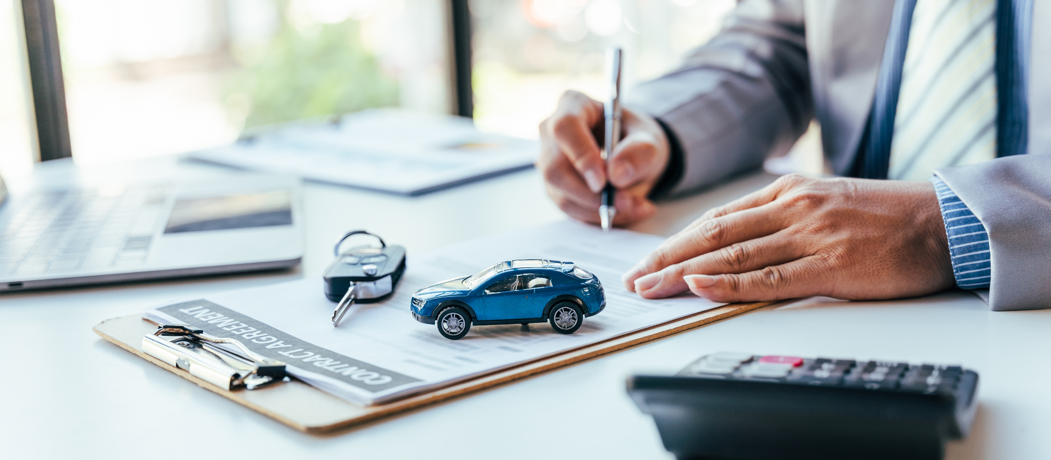 A person in a suit signs a contract on a desk with a miniature blue car, car keys, a laptop, and a calculator.