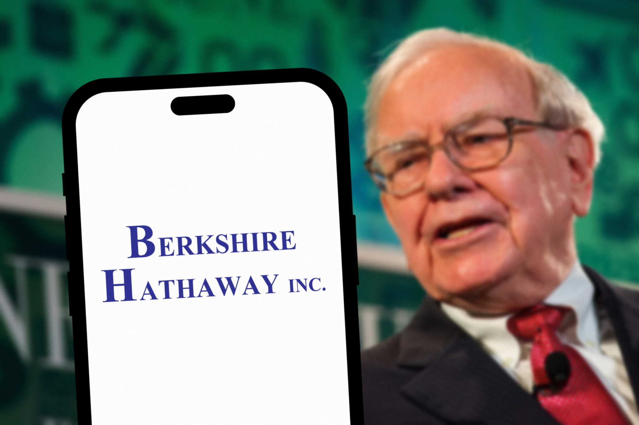 Warren Buffett speaking, with a smartphone showing the Berkshire Hathaway Inc. logo in the foreground, emphasizing strategy.