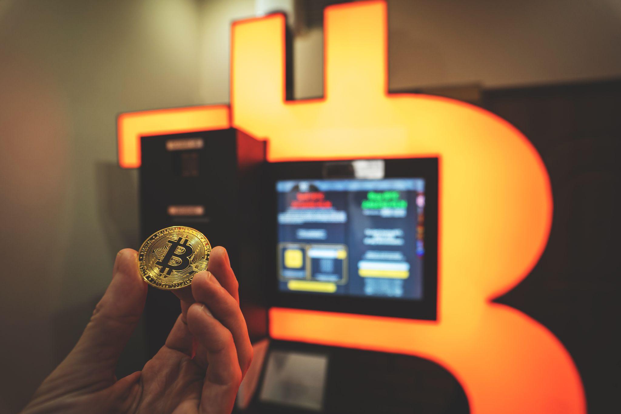 "Bitcoin gold coin held by hand next to an orange Bitcoin ATM for buying and selling BTC. Toned image with selective focus"