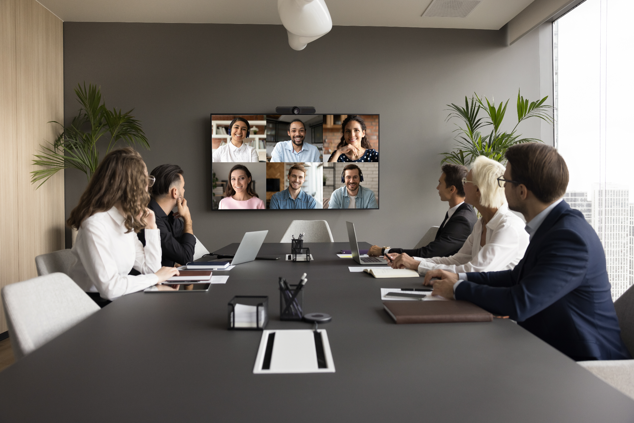 “Business professionals in a meeting room, engaged in a video conference with colleagues displayed on a large screen.”