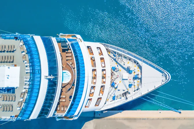 :”The nose of a luxury cruise ship docked near a busy pier, with clear blue water and a vibrant port in the background”.