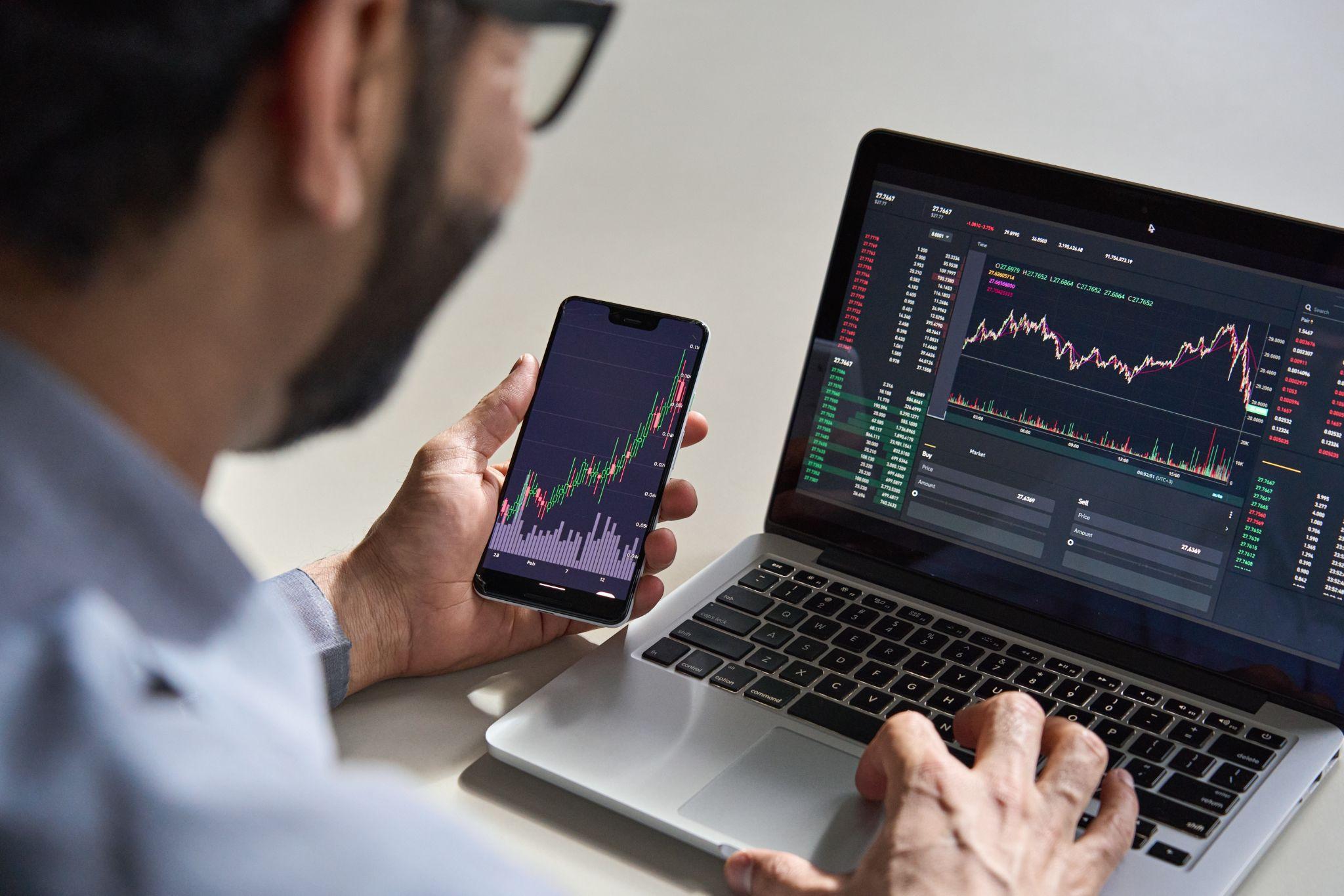 Businessman analyzing cryptocurrency market data on phone app and laptop, viewing trading charts and index graphs.