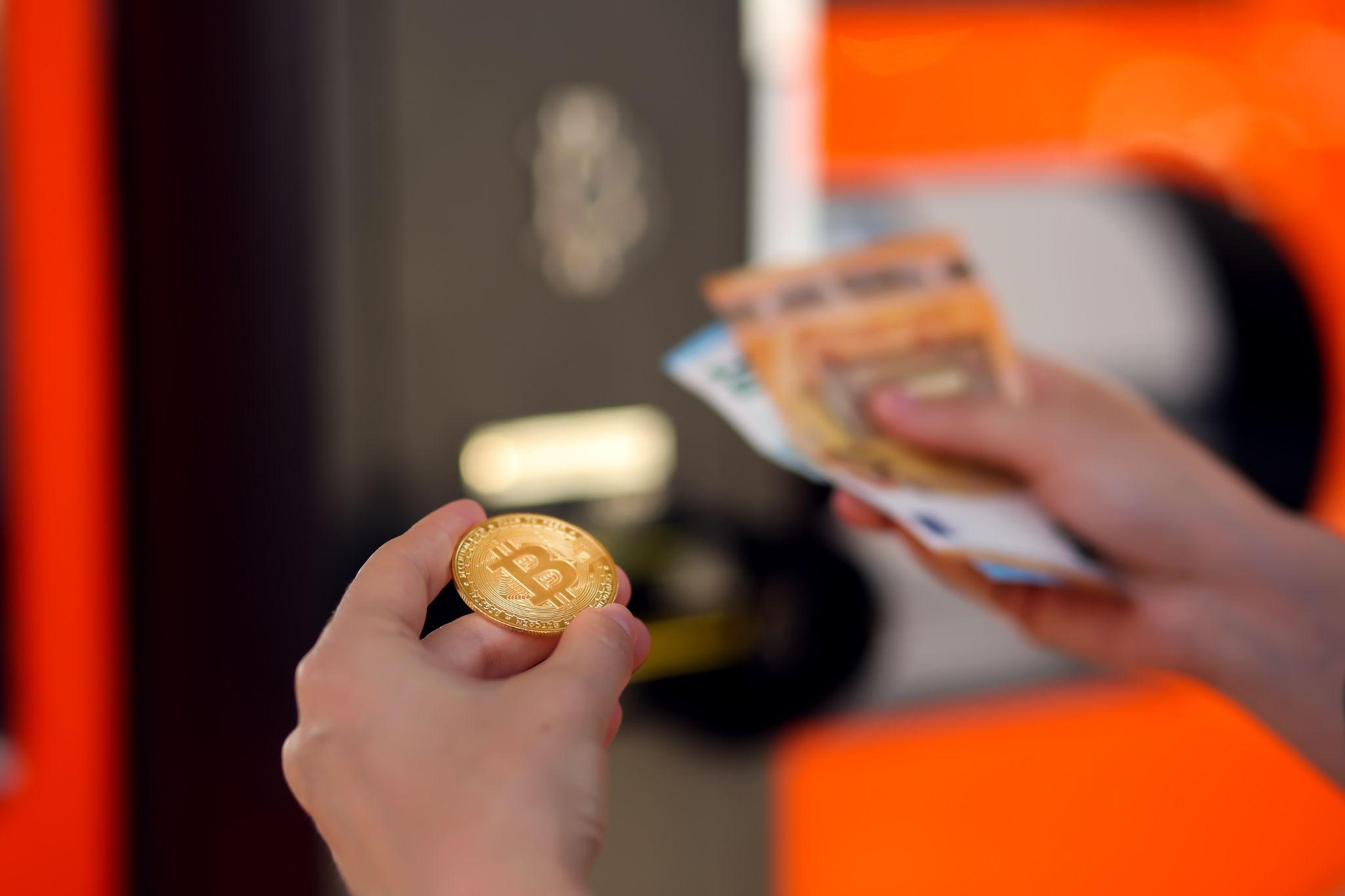 "Bitcoin gold coin held by hand next to an orange Bitcoin ATM for buying and selling BTC. Toned image with selective focus"