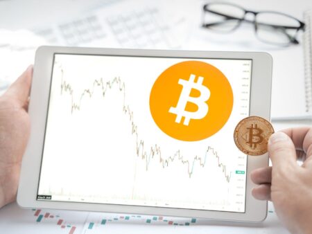 Tablet showing Bitcoin logo and stock price chart on trading platform. Blurred background with cryptocurrency trading tools