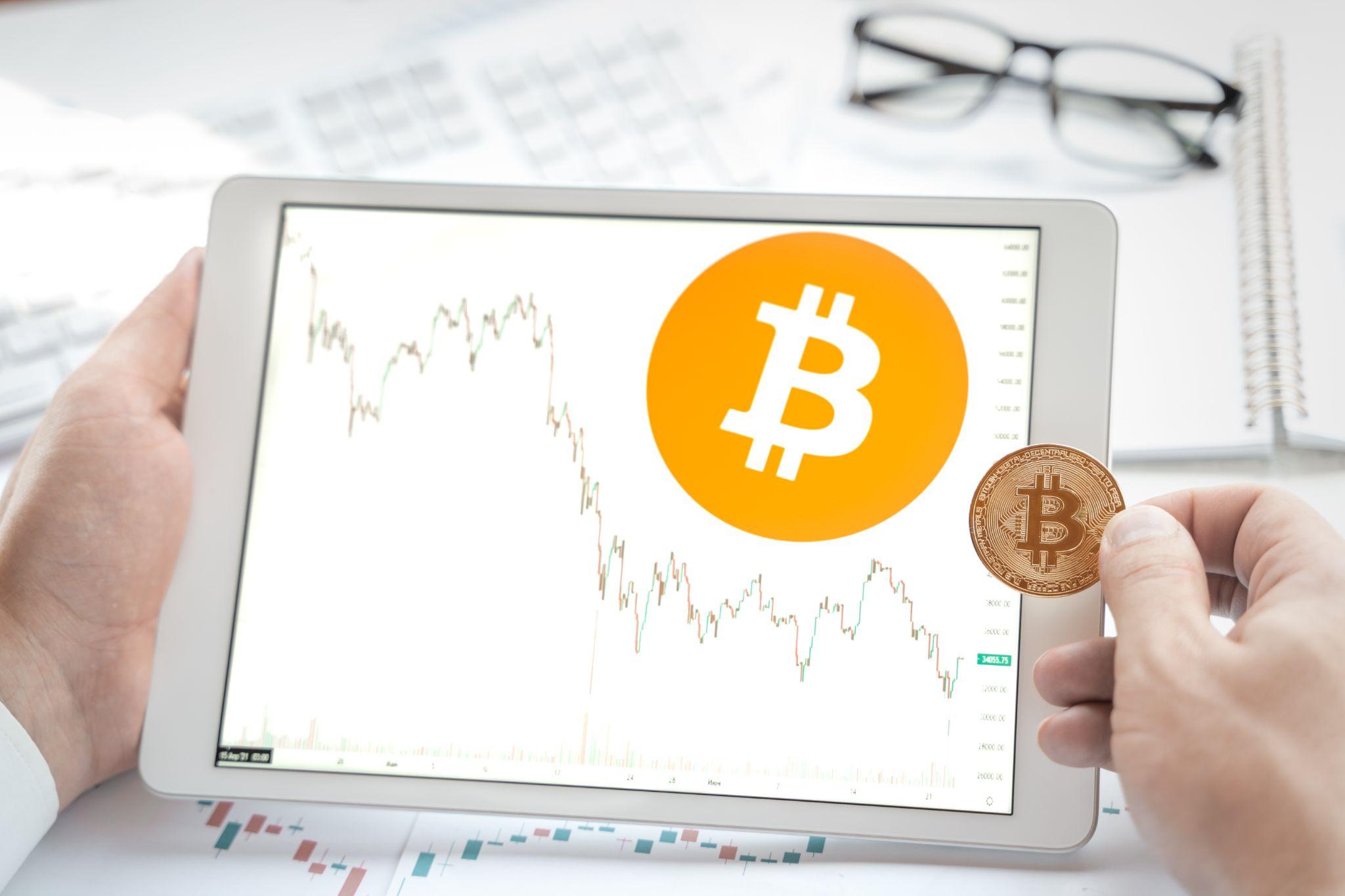 Tablet showing Bitcoin logo and stock price chart on trading platform. Blurred background with cryptocurrency trading tools