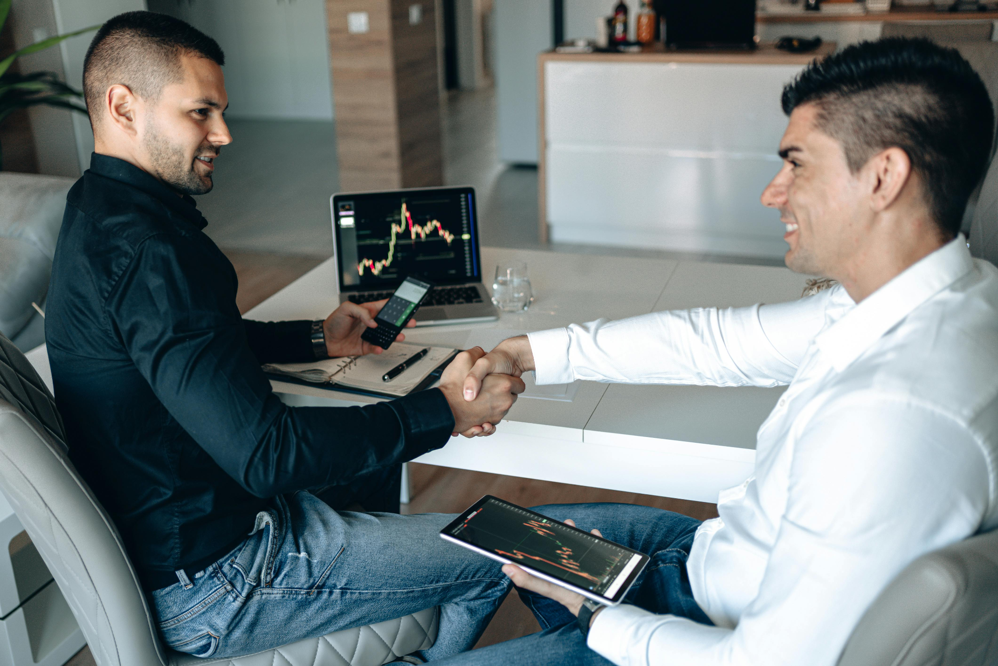 Two businessmen shaking hands over a successful trade, with trading charts displayed on a laptop and a tablet on the table.