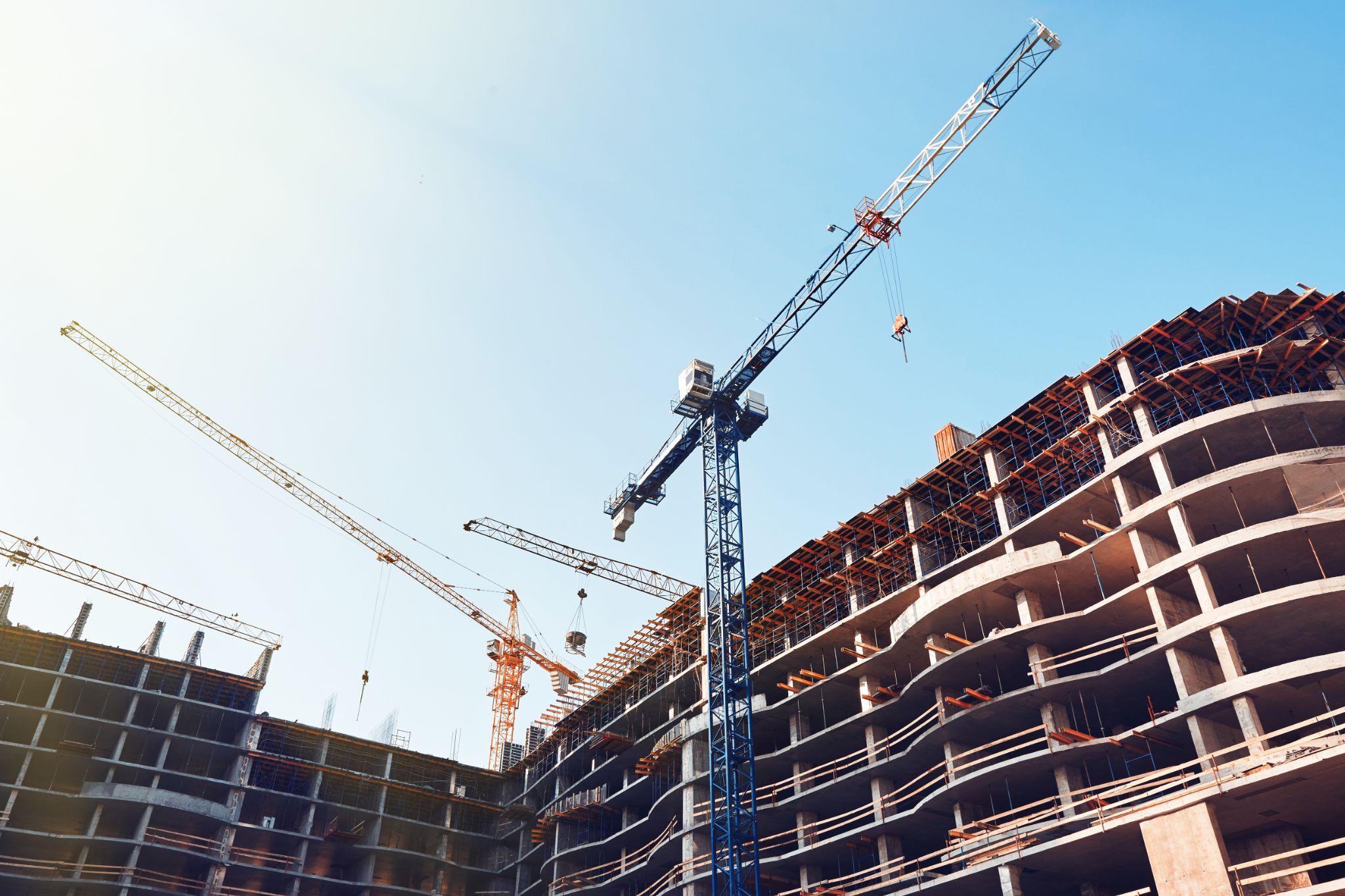 High building under construction. Side with cranes against blue sky with sun glare.