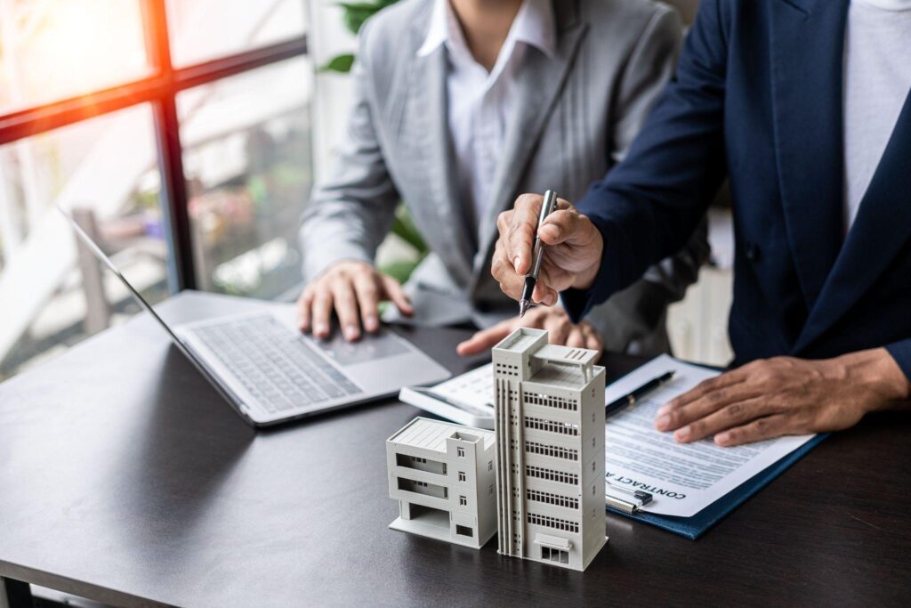 Agents offer housing contracts. Businessman holds small house model with property insurance at home sales office table